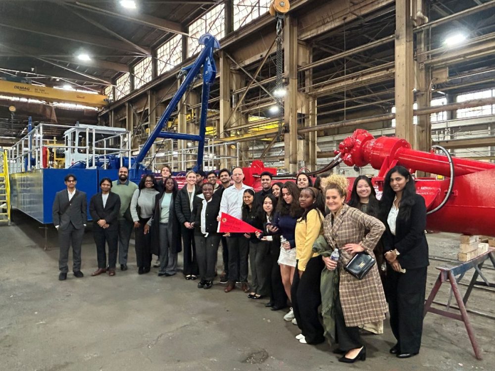 a diverse group of young adults poses for a group photo in front of a dredge in a warehouse
